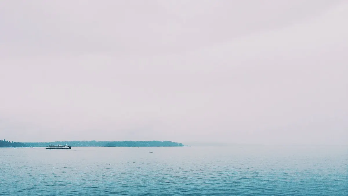 Mer calme sous un ciel pâle et brumeux, un ferry glissant au loin devant une île boisée